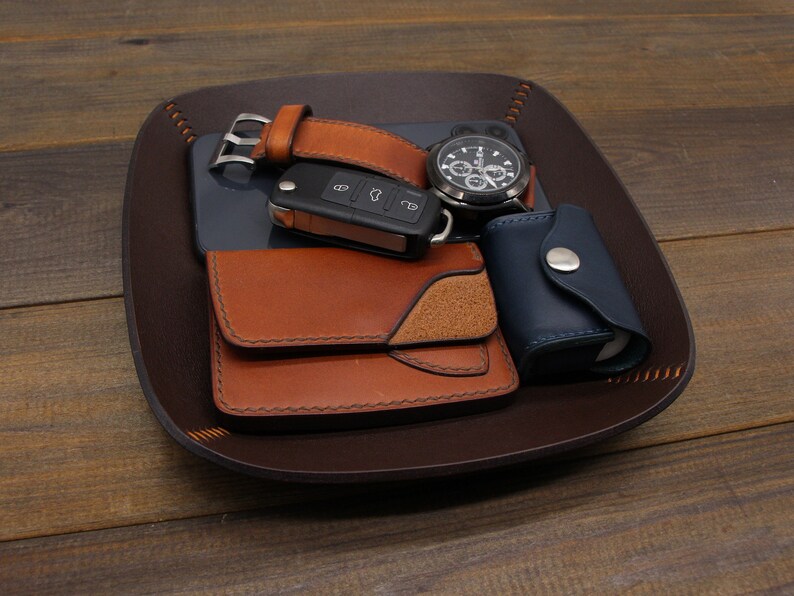 Puede incluir: Una bandeja de cuero marr&oacute;n con un reloj, una llave, una cartera y una peque&ntilde;a bolsa azul. La bandeja est&aacute; hecha de cuero y tiene un borde cosido.