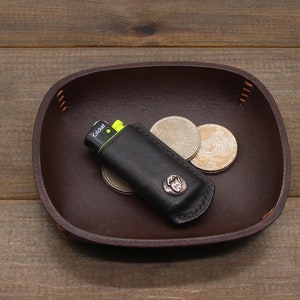 May include: A brown leather tray with a black leather lighter case and three silver coins. The lighter case has the word "Cricket" printed on it.