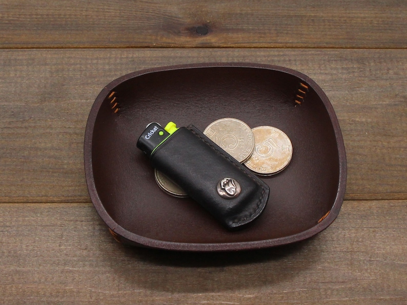 May include: A brown leather tray with a black leather lighter case and three silver coins. The lighter case has the word "Cricket" printed on it.