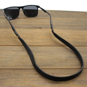 May include: Black sunglasses with a black leather strap. The sunglasses have a rectangular frame and dark lenses. The strap is attached to the temples of the sunglasses and is designed to be worn around the neck.