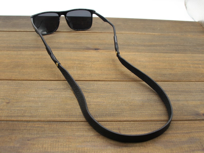 May include: Black sunglasses with a black leather strap. The sunglasses have a rectangular frame and dark lenses. The strap is attached to the temples of the sunglasses and is designed to be worn around the neck.