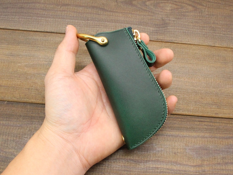 Puede incluir: Un llavero de cuero verde oscuro con un anillo de lat&oacute;n y una cremallera. El llavero est&aacute; en una mano.