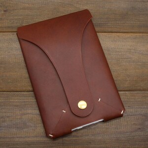 May include: A brown leather envelope-style case with a flap closure and a brass button. The case has white stitching around the edges.