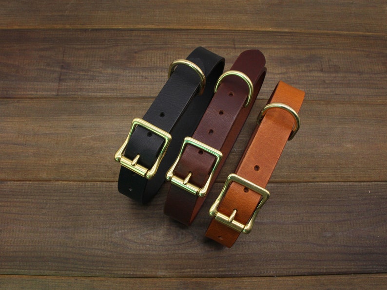 May include: Three leather dog collars in black, brown, and tan with brass buckles. The collars are arranged in a row on a wooden surface.