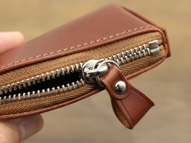 May include: A brown leather zippered wallet with a silver zipper pull. The wallet is closed and the zipper is pulled up.