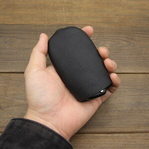 Puede incluir: Una funda de cuero negro con una superficie texturizada. La funda tiene una forma redondeada y una peque&ntilde;a abertura en un lado.