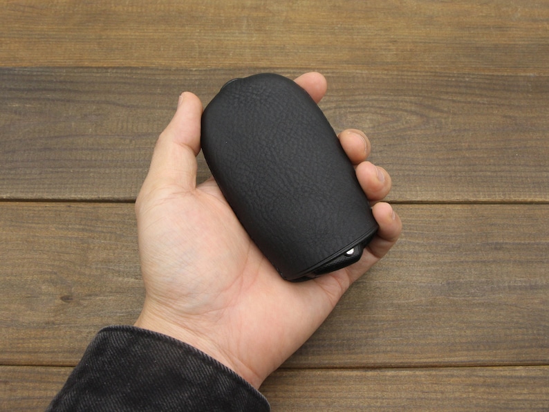 Puede incluir: Una funda de cuero negro con una superficie texturizada. La funda tiene una forma redondeada y una peque&ntilde;a abertura en un lado.