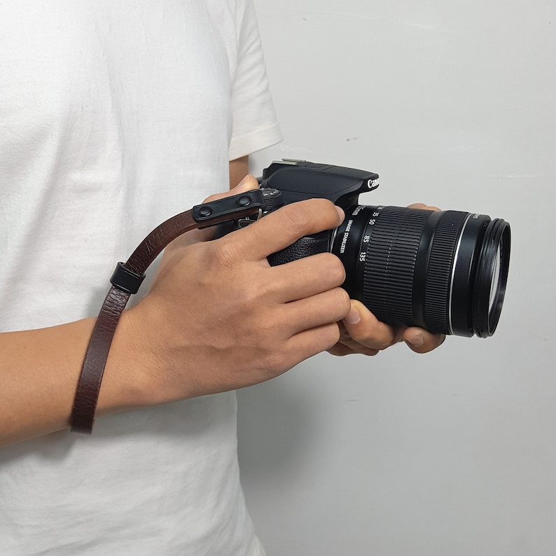 May include: A brown leather camera wrist strap with a metal buckle. The strap is attached to a black DSLR camera with a lens.
