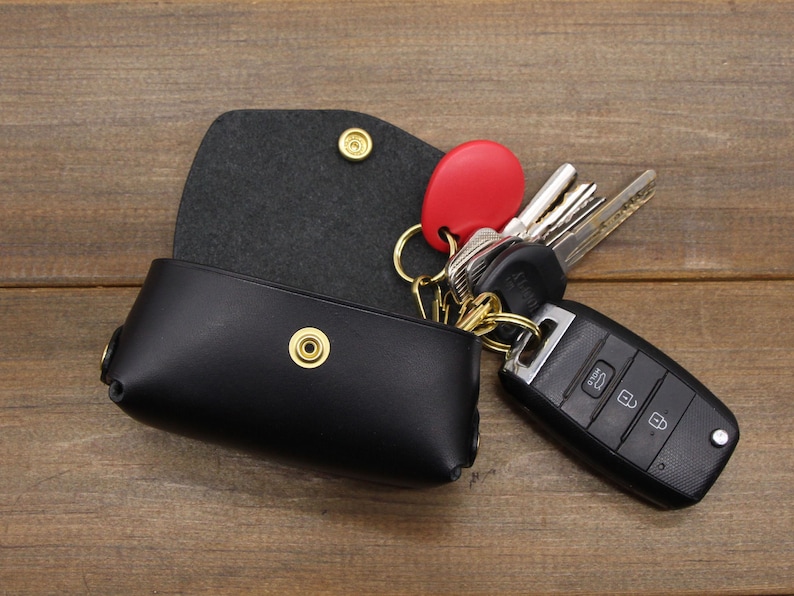 May include: Black leather key holder with a snap closure and a gold metal snap. The key holder is open and a set of keys with a red fob is attached to a gold metal key ring.