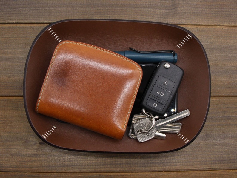 May include: A brown leather wallet, a car key with a key chain, and a pen are in a brown leather valet tray.