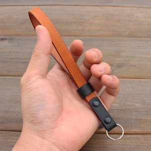 Pu&ograve; includere: Un cinturino per fotocamera in pelle marrone con dettagli in pelle nera e un portachiavi argentato.