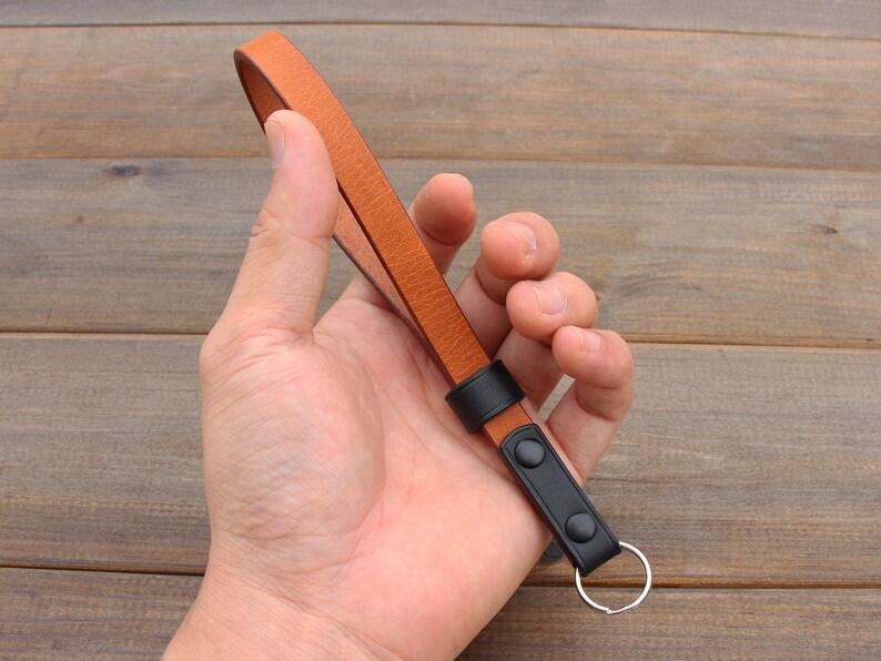 Pu&ograve; includere: Un cinturino per fotocamera in pelle marrone con dettagli in pelle nera e un portachiavi argentato.