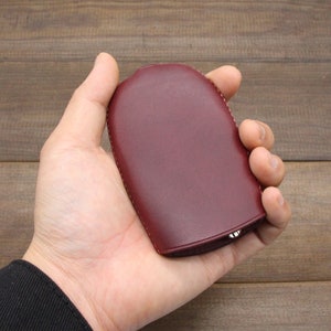May include: A burgundy leather key case with a rounded shape. The case is held in a hand, showcasing its smooth texture and stitched edges. The key case appears to be closed, suggesting it securely holds keys.