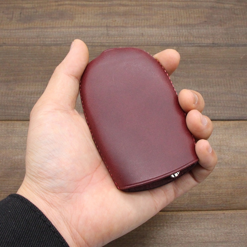 May include: A burgundy leather key case with a rounded shape. The case is held in a hand, showcasing its smooth texture and stitched edges. The key case appears to be closed, suggesting it securely holds keys.