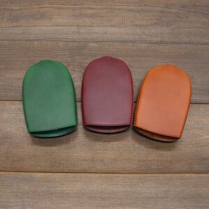 May include: Three leather key cases in green, burgundy, and tan. Each case has a rounded top and a stitched edge. The cases are arranged on a wooden surface.
