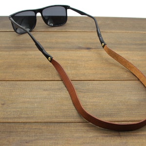 May include: Black sunglasses with a brown leather strap. The strap is attached to the glasses' arms with small gold-colored rivets. The sunglasses are resting on a wooden surface.