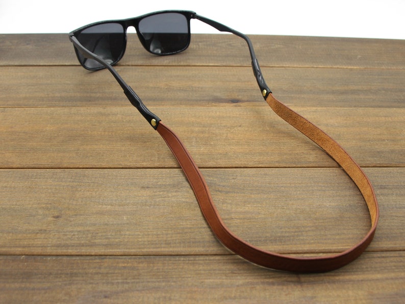 May include: Black sunglasses with a brown leather strap. The strap is attached to the glasses' arms with small gold-colored rivets. The sunglasses are resting on a wooden surface.