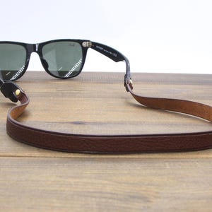 May include: Black sunglasses with green lenses and a brown leather strap. The strap is attached to the glasses and is designed to be worn around the neck. The sunglasses are resting on a wooden surface.