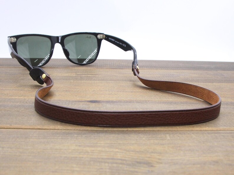 May include: Black sunglasses with green lenses and a brown leather strap. The strap is attached to the glasses and is designed to be worn around the neck. The sunglasses are resting on a wooden surface.