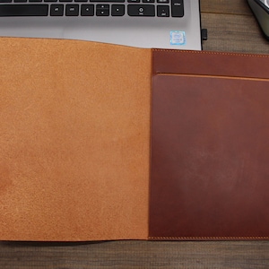 May include: A brown leather folder with a flap and a pocket on the front. The folder is open and the inside is lined with a light brown leather.