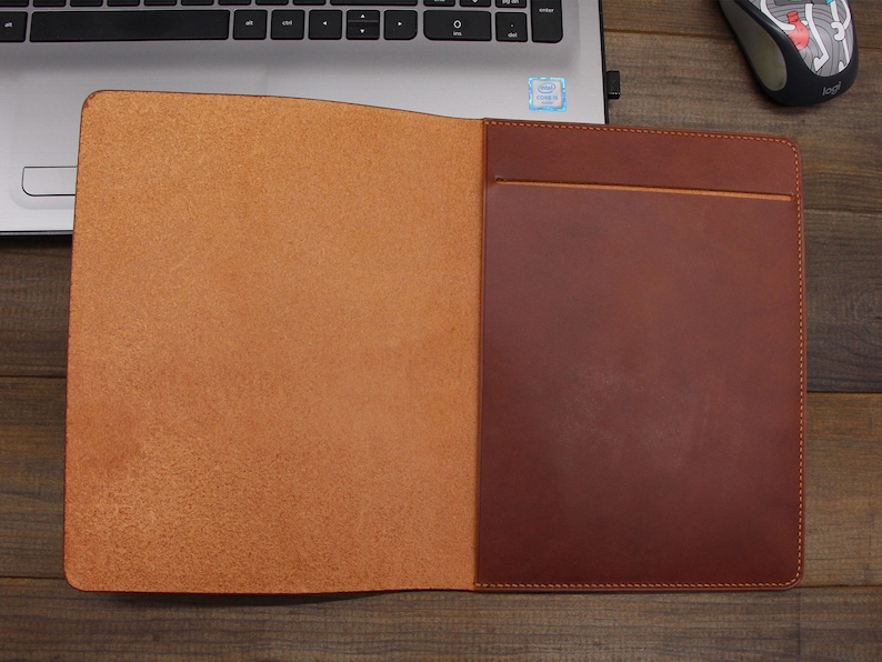 May include: A brown leather folder with a flap and a pocket on the front. The folder is open and the inside is lined with a light brown leather.