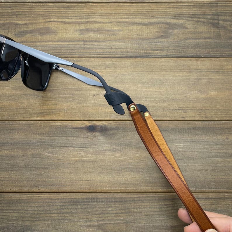 May include: Black sunglasses with silver frames are attached to a brown leather eyeglass strap. The strap is made of two strips of leather, held together with gold-colored rivets. The background is a wooden surface.