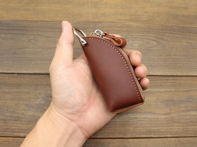 May include: A brown leather key chain with a zipper closure and a silver ring. The key chain is held in a hand.