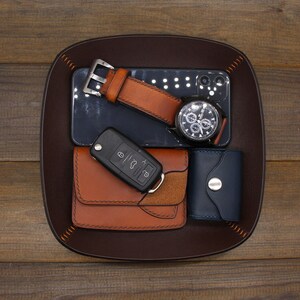 Puede incluir: Una bandeja de valet de cuero marr&oacute;n con un reloj de pulsera de cuero marr&oacute;n, un tel&eacute;fono inteligente negro, una cartera de cuero marr&oacute;n, una llave de coche y un tarjetero de cuero azul.