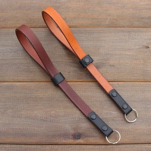 Pu&ograve; includere: Due cinturini per fotocamera in pelle con accessori in metallo nero e un anello argentato. Un cinturino &egrave; marrone e l'altro &egrave; arancione.