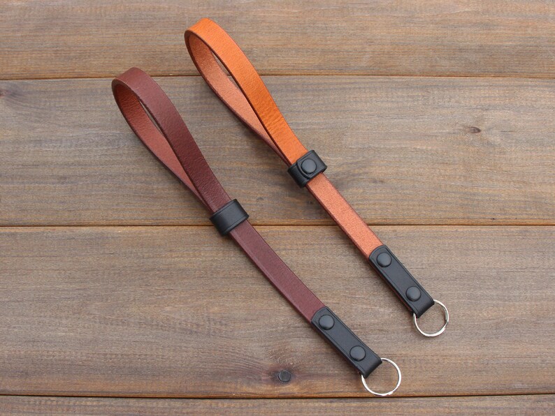 Pu&ograve; includere: Due cinturini per fotocamera in pelle con accessori in metallo nero e un anello argentato. Un cinturino &egrave; marrone e l'altro &egrave; arancione.