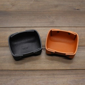 Puede incluir: Dos bandejas de cuero, una negra y otra marr&oacute;n, con forma cuadrada y esquinas redondeadas. La bandeja negra tiene la palabra "BLV" grabada en la parte inferior.