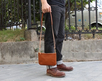 Può includere: Una borsa a tracolla in pelle marrone con una lunga tracolla. La borsa è indossata da una persona che indossa jeans blu e stivali marroni.