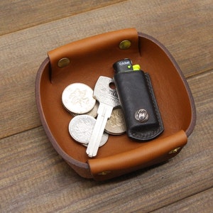 May include: A brown leather tray with a square shape and rounded edges. The tray has four brass rivets around the rim. The tray contains a key, a lighter, and several coins.