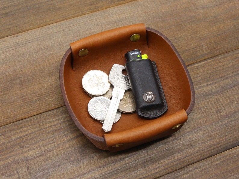 May include: A brown leather tray with a square shape and rounded edges. The tray has four brass rivets around the rim. The tray contains a key, a lighter, and several coins.