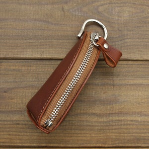 May include: A brown leather key chain with a zipper closure and a silver metal ring. The key chain is on a wooden surface.