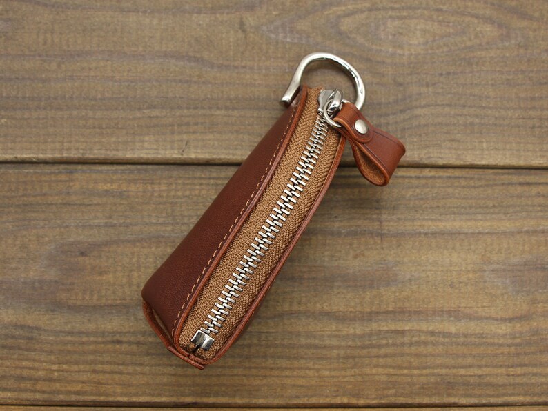 May include: A brown leather key chain with a zipper closure and a silver metal ring. The key chain is on a wooden surface.