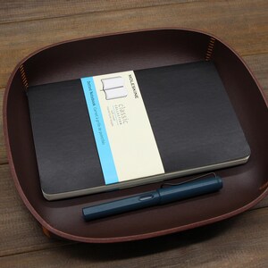 May include: A brown leather tray with a black notebook and a blue pen. The notebook has the text "Classic Collection" and "Ruled Notebook Cahier &agrave; lignes de papier" on the cover.