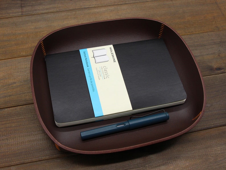 May include: A brown leather tray with a black notebook and a blue pen. The notebook has the text "Classic Collection" and "Ruled Notebook Cahier &agrave; lignes de papier" on the cover.
