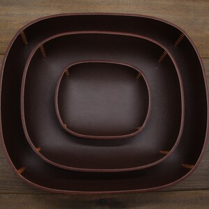 May include: Three brown leather trays of varying sizes, nested inside each other. The trays have a rounded, oval shape and are stitched together with brown thread.