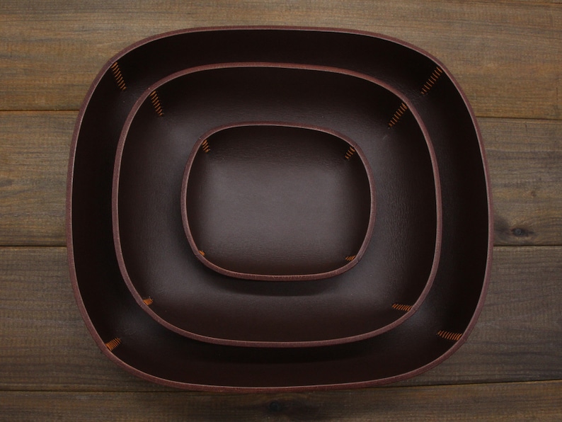 May include: Three brown leather trays of varying sizes, nested inside each other. The trays have a rounded, oval shape and are stitched together with brown thread.