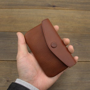 Puede incluir: Una cartera de cuero marr&oacute;n con cierre de solapa y bot&oacute;n. La cartera se sostiene en una mano.