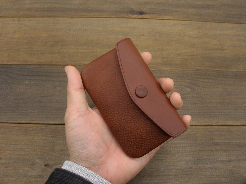 Puede incluir: Una cartera de cuero marr&oacute;n con cierre de solapa y bot&oacute;n. La cartera se sostiene en una mano.