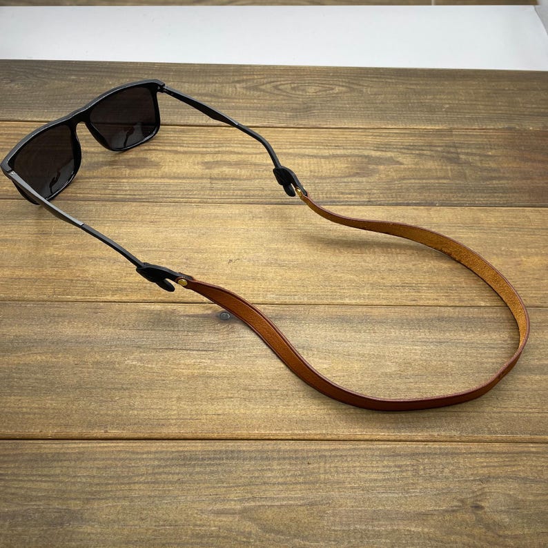 May include: Black sunglasses with a brown leather strap. The sunglasses have a rectangular frame and dark lenses. The strap is attached to the arms of the glasses with black connectors and is made of a smooth, light brown leather.