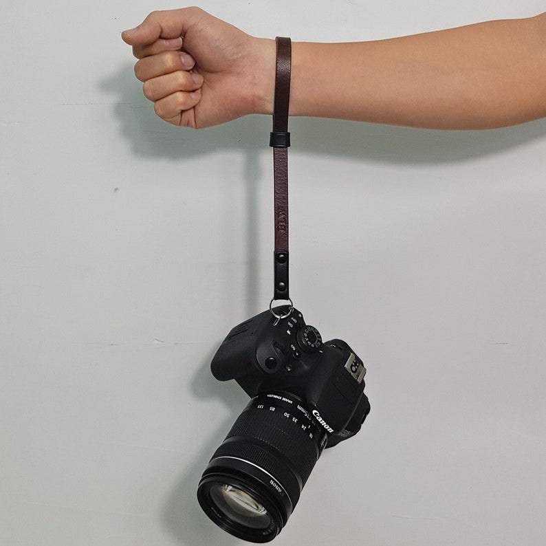 May include: A brown leather camera strap with a black metal clasp attached to a black Canon DSLR camera with a lens.