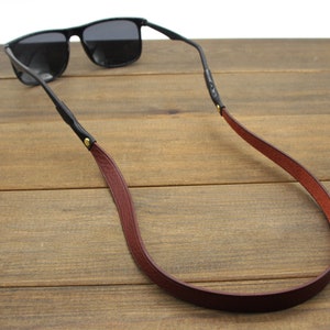 May include: Black sunglasses with a burgundy leather strap. The strap is attached to the glasses with black leather and gold-colored rivets. The sunglasses are resting on a wooden surface.