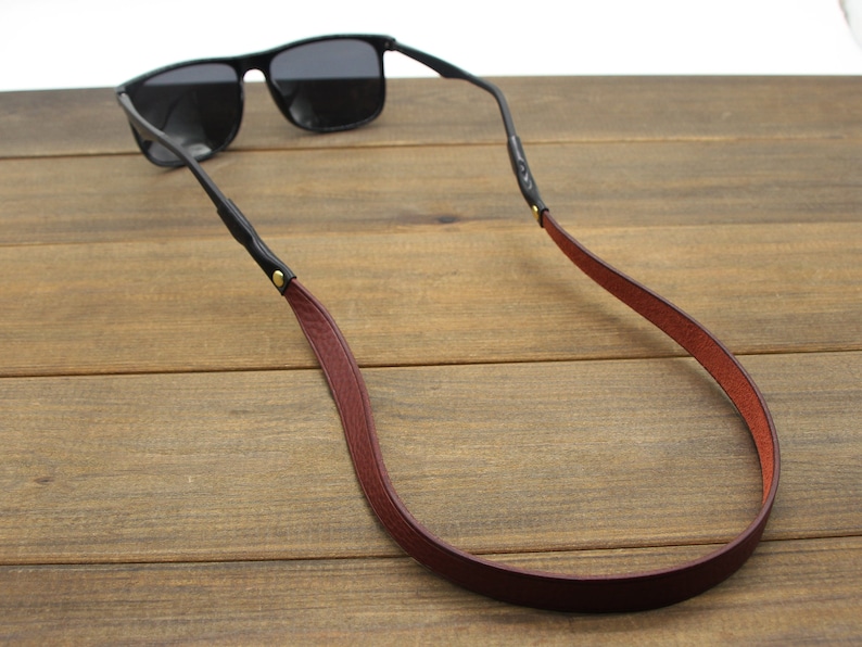 May include: Black sunglasses with a burgundy leather strap. The strap is attached to the glasses with black leather and gold-colored rivets. The sunglasses are resting on a wooden surface.