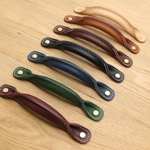 Könnte beinhalten: Eine Auswahl an Leder-Schrankgriffen in verschiedenen Farben: Beige, Braun, Marineblau, Grün und Burgunderrot. Jeder Griff hat weiße Nähte und messingfarbene Nieten, angeordnet auf einer hellen Holzoberfläche.