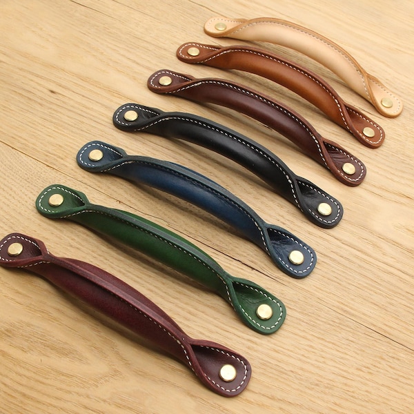 May include: An assortment of leather cabinet handles in various colors, including tan, brown, navy, green, and burgundy. Each handle features white stitching and brass-colored rivets, arranged on a light wood surface.