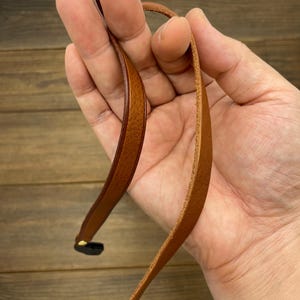 May include: Close-up of a brown leather strap with a black end piece. The strap is held in a hand against a wooden background. The leather appears to be new and unworn.