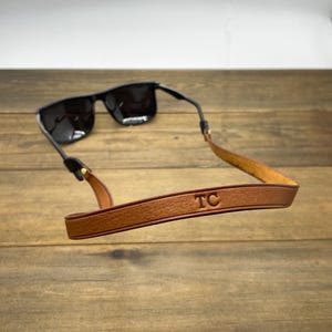 May include: Black sunglasses with a brown leather strap. The strap has the initials "TC" embossed on it. The sunglasses are resting on a wooden surface.